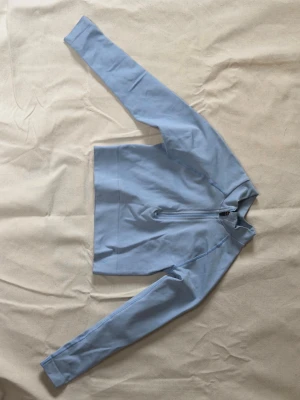 Ljusblå träningströja från TALA - Ljusblå långärmad träningströja från TALA med halv dragkedja framtill, kort i midjan. Tillverkad i en mjuk och stretchig bomullsblandning, perfekt för träning eller sportiga aktiviteter. Snygg och enkel design med diskreta sömmar.