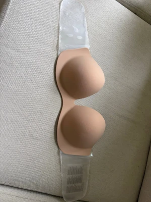 Beige självhäftande bandeau-bh - Självhäftande bandeau-bh i beige färg, perfekt för axelbandslösa eller rygglösa plagg. Bh:n har slät form och silikon på insidan för att sitta på plats. Levereras i originalförpackning.Har endast provats, inte använts men kan såklart ha fastnat lite damm på självhäftningen. 