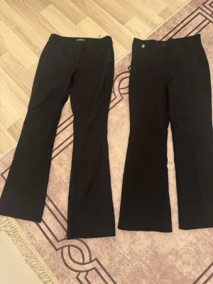 Svarta kostymbyxor med utsvängda ben - Två par svarta kostymbyxor med utsvängda ben och klassisk passform. Ena paret har diskret metallogo vid fickan och båda har hällor för skärp. Perfekta till en dressad look och passar till skjorta eller kavaj.