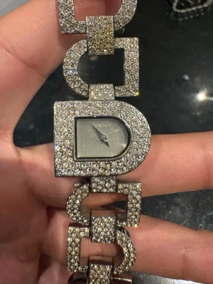 Glittrig silverklocka från DKNY - Säljer en unik och glittrig armbandsklocka från DKNY i silverfärg. Klockan har ett geometriskt armband täckt av gnistrande stenar och en rektangulär urtavla med DKNY-logga. Perfekt för dig som vill sticka ut med accessoarer som verkligen syns.