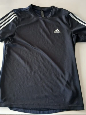 Mörkblå Adidas t-shirt med ränder - Mörkblå t-shirt från Adidas med klassiska vita ränder på axlarna och Adidas-logga på bröstet. T-shirten har rund hals och är kortärmad, perfekt för en sportig stil. Materialet är mjukt och känns bekvämt mot huden.