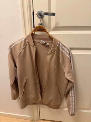 Beige Adidas ziptröja med ränder - Snygg beige ziptröja från Adidas med klassiska vita ränder längs ärmarna och diskret logga på bröstet. Tröjan har dragkedja framtill, två fickor med dragkedja och ribbade muddar. Perfekt för en sportig och avslappnad stil.