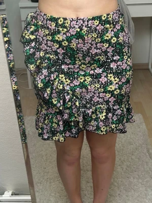 Blommig kortkjol med volang - Säljer en färgglad kortkjol med blommigt mönster i svart, grönt, gult, rosa och lila. Kjolen har volangdetaljer och en omlott-look med knyt på sidan. Perfekt för dig som gillar att sticka ut med din stil!