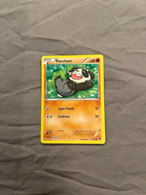 Pokémonkort Pancham - Säljer ett Pokémonkort med karaktären Pancham. Kortet har en färgglad illustration med gröna och gula detaljer, och text i svart och orange. Kortet är tillverkat i laminerad papp och har attackerna Light Punch och Confront.