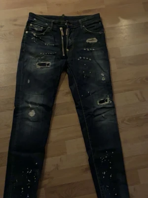  Dsquared2 Jens  - Snygga mörkblå jeans från Dsquared2 med slitningar, hål och färgstänk för en riktigt edgy look. Klassisk femficksmodell med silverfärgad dragkedja och Dsquared2-logga vid gylfen. Perfekta för dig som gillar streetstyle.
