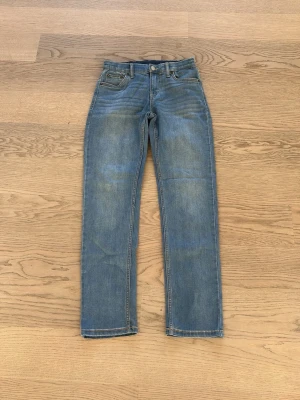 Blå raka jeans från Levi's - Klassiska blå jeans från Levi's med rak passform och fem fickor. Jeansen har en ljus tvätt och synliga slitningar vid knän och lår. Tillverkade i bomull med Levi's ikoniska läderpatch bak i midjan och röda etiketten på bakfickan.