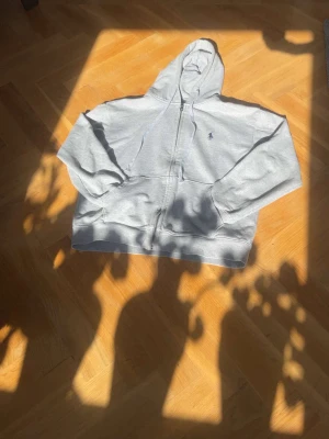 Grå zip-hoodie från Polo Ralph Lauren - Snygg grå hoodie med dragkedja från Polo Ralph Lauren. Klassisk design med huva, två fickor fram och liten broderad logga på bröstet. Perfekt för en chill och stilren look. Mjuk bomullskänsla och relaxed fit.
