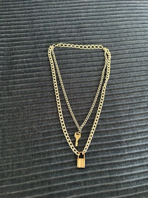Silver halsband med lås och nyckel - Dubbelkedjat halsband i silver metall med coolt hängsmycke i form av ett hänglås och en liten nyckel. Kedjorna har olika tjocklek för en trendig lager-på-lager-look. Perfekt accessoar för att lyfta din outfit.