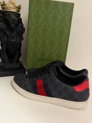 Gucci sneakers  - Snygga Gucci sneakers i mörkblå canvas med GG-mönster, klassiska röda och blå ränder på sidan samt metallicröd och metalliclila detalj på hälen. Låg modell med vita sulor och svarta skosnören. Perfekt för dig som vill sticka ut med exklusiva skor. Storlek uk 8 