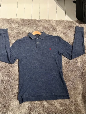 Blå långärmad Ralph Lauren piké  - Klassisk blå långärmad piké från Polo Ralph Lauren med krage och två knappar vid halsen. Perfekt för en stilren och avslappnad look.