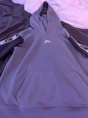 Grå Nike hoodie med logga - Snygg grå hoodie från Nike med vit logga på bröstet och svarta ärmar med vita Nike-band längs sidorna. Klassisk känguruficka framtill och huva. Perfekt för en sportig och avslappnad stil. PRISET KAN DISKUTERAS!
