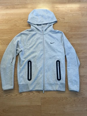 Nike NOCTA  - Snygg ljusgrå hoodie från Nike NOCTA med hel dragkedja framtill, svarta detaljer vid fickorna och liten Nike-logga på bröstet. Huva och tryck på ärmen samt diskreta tryck på ryggen. Perfekt för en sportig och stilren look.