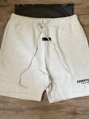 Gråa Essentials shorts med snörning - Snygga ljusgrå shorts från Fear of God Essentials med elastisk midja och snörning framtill. Mjuk bomullskvalitet och diskret logga på benet. Perfekta för en avslappnad och trendig stil.