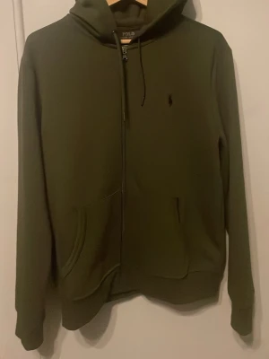 Olivgrön hoodie från Polo Ralph Lauren - Snygg olivgrön hoodie från Polo Ralph Lauren med dragkedja framtill och klassisk broderad logga på bröstet. Hoodien har känguruficka, justerbar huva med snörning och är tillverkad i mjukt material. Perfekt för en chill och stilren look. Storlek M