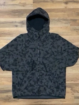 Svart camo hoodie från Nike - Snygg hoodie från Nike i svart och mörkgrått camo-mönster. Dragkedja framtill, två fickor och justerbar huva. Perfekt för en streetwear-look och passar till det mesta. Tillverkad i mjukt material för extra komfort.