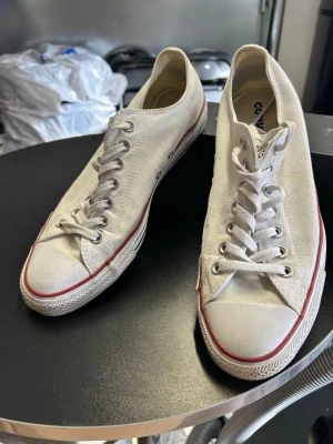 Vita Converse All Star låga sneakers - Klassiska vita Converse All Star sneakers med lågt skaft, vita snören och röd rand längs sulan. Skorna har canvasöverdel och gummisula, perfekt för en avslappnad och tidlös stil.