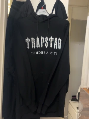  Trapstar hoodie - Svart hoodie från Trapstar med stort vitt tryck på bröstet där det står 'TRAPSTAR IT'S A SECRET'. Klassisk modell med huva och känguruficka. Perfekt för dig som gillar streetwear och vill sticka ut med en snygg logga. Back logga finns, köpt från trapstar 2024 men knappt använd. Strl S/M