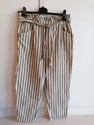 Randiga tygbyxor med knytskärp - Tygbyxor i beige med svarta och vita vertikala ränder. Byxorna har volangmidja, knytskärp och två framfickor. Avslappnad och croppad passform, perfekt för en chill och trendig stil.