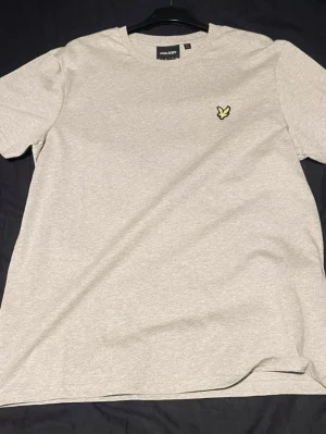 Ljusgrå t-shirt från Lyle & Scott - Snygg ljusgrå t-shirt från Lyle & Scott med klassisk passform och korta ärmar. T-shirten har en gul broderad logga på bröstet och är tillverkad i mjuk bomull. Perfekt till jeans eller shorts.