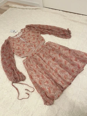 Mango sommar rose kortklänning med volang L - Supersöt kortklänning med volang och långa ärmar i ett blommigt och paisley-inspirerat mönster i rosa och beige toner. Klänningen har rund halsringning med liten öppning framtill och resår i midjan för en bekväm passform. Perfekt för vår och sommar!