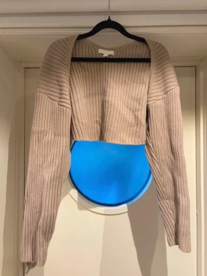 Beige ribbstickad bolero - Säljer en beige ribbstickad boleron med långa ärmar och öppen front. Tröjan har en modern, avslappnad passform och är perfekt att slänga över en topp. Materialet är mjukt och stretchigt, vilket gör den både bekväm och snygg.