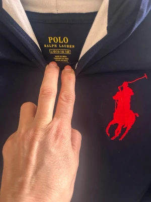 Marinblå hoodie från Polo Ralph Lauren - Snygg marinblå hoodie från Polo Ralph Lauren med stor röd logga på bröstet. Hoodien har huva med vit insida och långa ärmar. Perfekt för dig som gillar klassisk och sportig stil. Tillverkad i mjukt material för skön känsla.