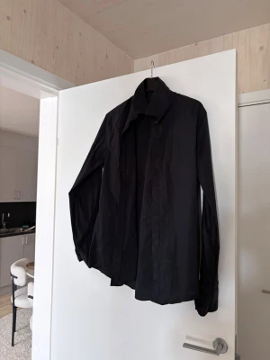 Svart klassisk skjorta i bomull - En stilren svart skjorta med lång ärm och klassisk krage. Skjortan är tillverkad i bomull och har en rak passform. Perfekt för dig som gillar en enkel och clean look.