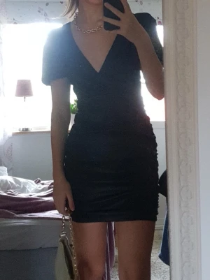 Svart klänning i satin - Svart kort omlottklänning i satin med v-ringning och korta puffärmar. Klänningen har en figurnära passform och rynkade detaljer längs sidorna.