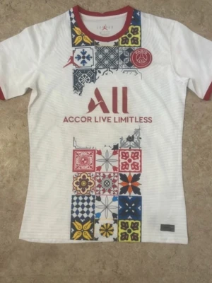 PSG x Jordan vit t-shirt med mönster - Vit t-shirt från Jordan x PSG med färgglada grafiska mönster och logga framtill. Röd krage och ärmslut, tryckt text och emblem. Perfekt för dig som gillar sportig och unik stil.