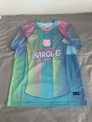 Nike x Karol G FC Barcelona tröja - Unik FC Barcelona tröja i samarbete med Karol G. Flerfärgad design i pastell med tryck fram och bak, inklusive 'Karol G' och 'Bichota' med hjärtmotiv och tecknad figur. Korta ärmar, sportig passform och tillverkad i ventilerande polyester.