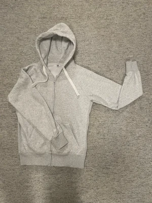 Ljusgrå hoodie med dragkedja L - Klassisk ljusgrå hoodie från Rushemerch i storlek L. Hoodien har dragkedja framtill, två fickor och justerbar huva med vita snören. Tillverkad i mjuk bomullsblandning som känns skön mot huden. Perfekt för en avslappnad och stilren look.