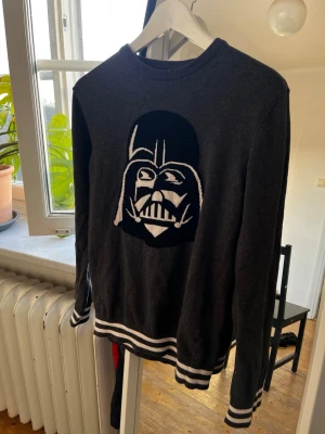 Mörkgrå stickad tröja med Darth Vader - Cool mörkgrå stickad tröja med stor Darth Vader-figur på bröstet. Tröjan har vita ränder vid ärmslut och nederkant samt rund halsringning. Perfekt för Star Wars-fans som vill sticka ut med sin stil. Köpt på comic con