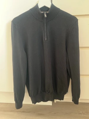 Half zip från Hugo Boss - Svart stickad tröja med hög krage och halv dragkedja framtill. Tröjan har långa ärmar, ribbstickade muddar och nederkant. Perfekt för lager-på-lager och enkel att matcha med olika outfits.