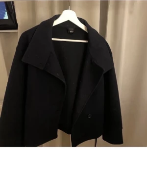 Svart kort ullrock med bred krage - Stilren svart kort kappa med bred krage och knäppning framtill. Enkel och snygg design som passar till många outfits. Köpt här på Plick men är i jätte bra skick!😊skriv om ni fundera på nåt!