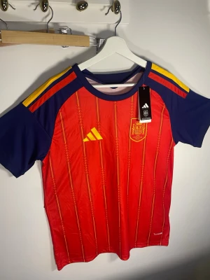Spanien landslags tröja Adidas  - Röd matchtröja från Adidas för Spaniens landslag med gula och blå detaljer. Tröjan har korta ärmar, rund hals och tunna vertikala ränder i gult. Broderad landslagssköld och Adidas-logga på bröstet. Tillverkad i lätt och ventilerande material.