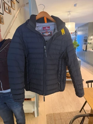 Svart pufferjacka Parajumpers - Snygg svart pufferjacka från Parajumpers med klassisk gul detalj vid kragen och dragkedja framtill. Jackan har huva, två sidofickor med dragkedja och quiltad design. Materialet är lätt och smidigt. Ny priss 5600kr