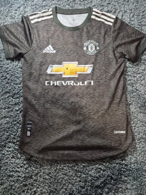 Manchester United bortatröja Adidas S - Säljer en Manchester United bortatröja från Adidas i storlek S. Tröjan har ett mörkgrönt och svart zebramönster, klassisk rund hals och korta ärmar. Materialet är lätt och andas, med Heat.RDY-teknologi. Loggor från Adidas, Manchester United och Chevrolet på bröstet.