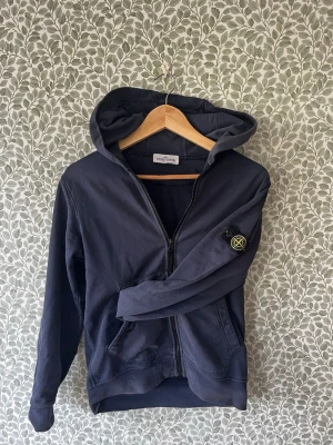 Mörkblå hoodie från Stone Island - Tjena! Säljer nu min mörkblå zip hoodie från Stone Island med anledning att den helt enkelt blivit för liten. Den har dragkedja, huva, två framfickor och den klassiska logotyp-patchen på ärmen. Har några små slitningar, se bild 4, men är annars i gott skick. Hör av dig angående frågor!