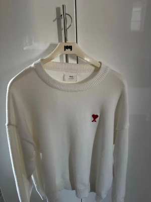 Vit stickad tröja från Ami Paris - Vit stickad tröja från Ami Paris med rund halsringning och ribbade muddar. På bröstet finns Ami Paris ikoniska röda hjärta med bokstaven A broderat. Enkel och stilren design som passar perfekt till många outfits. 