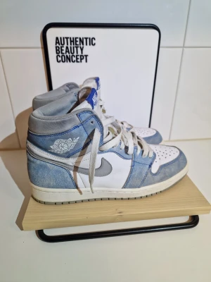 Nike Air Jordan 1 Retro High OG 'Hyper Royal  - Säljer ett par Nike Air Jordan 1 Retro High OG 'Hyper Royals neakers i blått och vitt med grå detaljer. Skorna har klassisk hög siluett, vit tåbox med perforeringar, blå mockadetaljer och vita skosnören. Swoosh-logga i grått på sidan och Air Jordan-märke på ankeln.