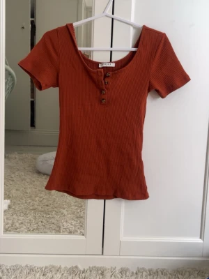 Vintage Rostbrun ribbad topp från Pieces - Säljer en ribbad rostbrun topp från Pieces i storlek XS. Toppen har korta ärmar, rund halsringning och fyra dekorativa knappar framtill. Materialet är mjukt och stretchigt, perfekt för en avslappnad look.