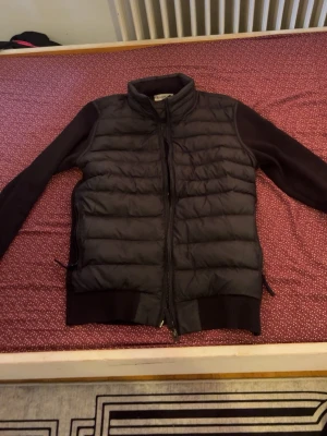 Svart dunväst från Moncler - Snygg svart dunväst från Moncler med quiltad front och mjuka stickade ärmar. Västen har hög krage, dragkedja framtill och Moncler-logga på ärmen. Perfekt för lager-på-lager och ger en clean look. Materialet är en mix av dun och bomull.