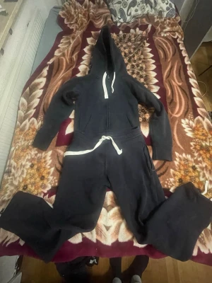 Mörkblå jumpsuit med huva XS - Mysig mörkblå jumpsuit från R i storlek XS. Har dragkedja framtill, stor huva och vita snören i midjan och vid huvan. Tillverkad i mjukt sweatshirtmaterial, perfekt för chill dagar hemma eller när du vill vara extra bekväm.