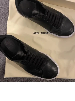 Svarta Axel Arigato sneakers i läder - Stilrena svarta sneakers från Axel Arigato i mjukt läder med klassisk låg profil och vita sulor. Skorna har svarta skosnören och minimalistisk design som passar till det mesta. Perfekta för dig som gillar en clean och modern look.