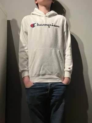 Vit Champion hoodie med broderad logga - Säljer en vit hoodie från Champion med broderad logga i blått och rött på bröstet. Hoodien har en stor ficka framtill, ribbade muddar och huva. Perfekt för dig som gillar streetwear och vill ha något bekvämt och snyggt.