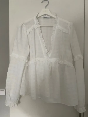 Vit volangblus med v-ringning - Söt vit blus med djup v-ringning och volangdetaljer längs halsringning, axlar och ärmslut. Lätt och luftig struktur, perfekt för en trendig och romantisk look. Långa ärmar med diskreta volanger ger en extra touch.