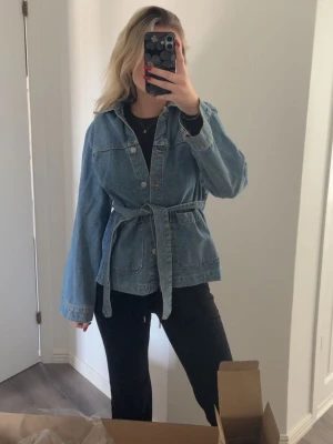 Blå jeansjacka med knytskärp - Snygg blå jeansjacka med knytskärp i midjan och stora fickor framtill. Klassisk modell med knappar och avslappnad passform. Perfekt att slänga över en hoodie eller t-shirt för en cool look.