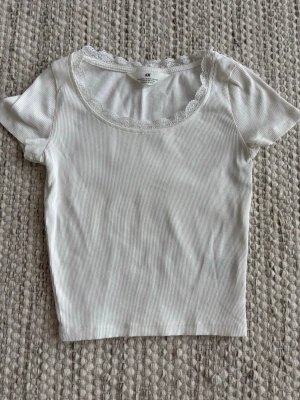 Vit ribbad topp med spets från H&M - Supersöt vit ribbad topp från H&M med korta ärmar och rund halsringning dekorerad med blommig spets. Perfekt att styla med jeans eller kjol. 