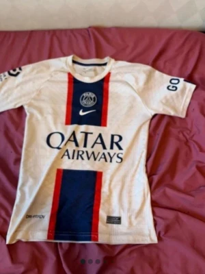 PSG Mbappé matchtröja Nike vit - Säljer en vit PSG matchtröja från Nike med röda och blå detaljer. Framsidan har Qatar Airways-logga och PSG-emblem, baksidan har Mbappé och nummer 7 tryckt. Korta ärmar och sportig passform, perfekt för fotbollsträning eller support.