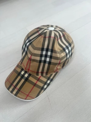 Beige keps med rutmönster - Snygg keps i klassiskt beige rutmönster med svarta, vita och röda linjer. Kepsen har böjd skärm och justerbart spänne bak med vit detalj. Perfekt accessoar för en trendig look.